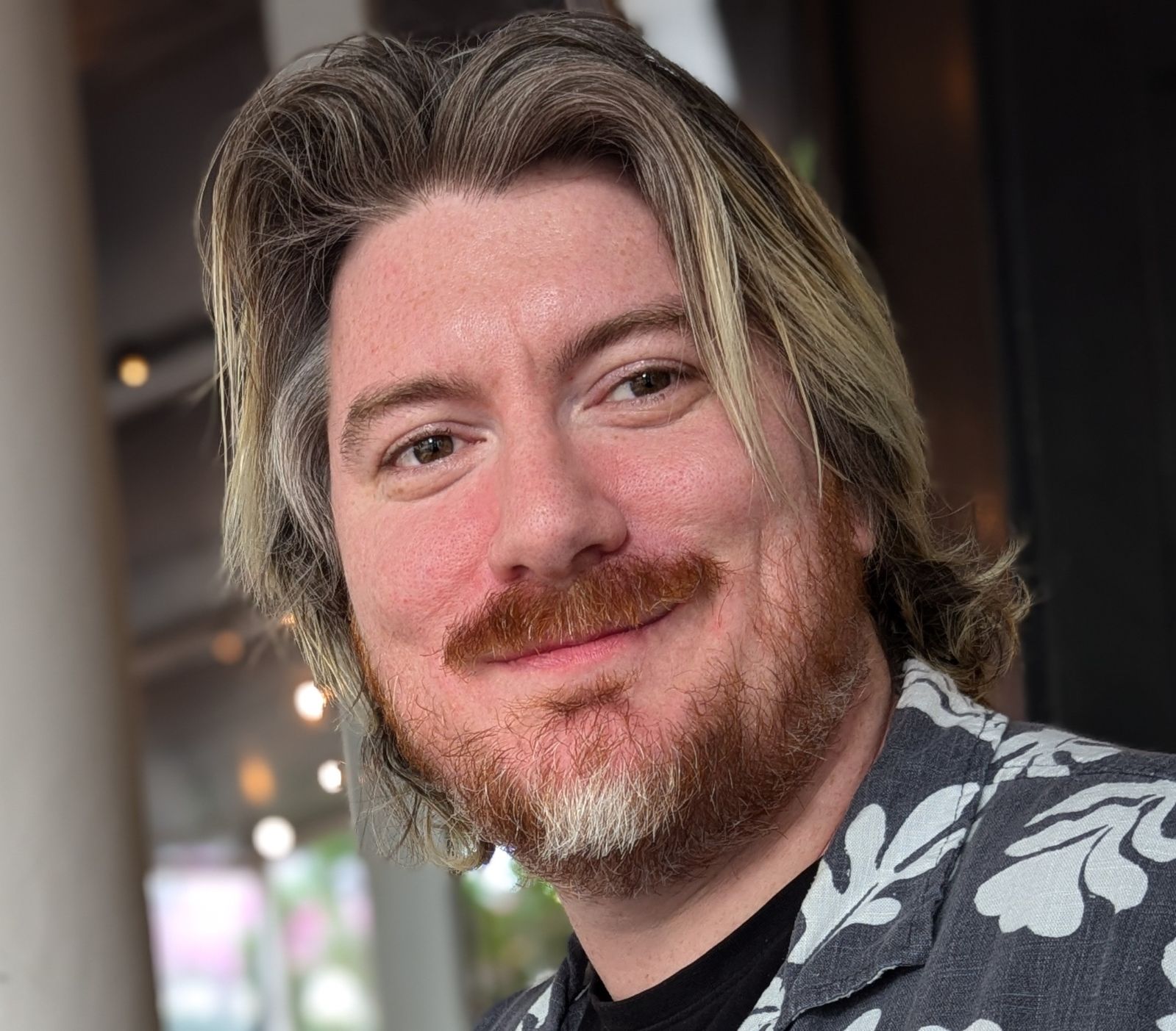 An image of Zigarot, wearing a black and white floral shirt, sporting ear-length long hair, and smiling at the camera. Beard and moustache is red and grey, hair is grey.